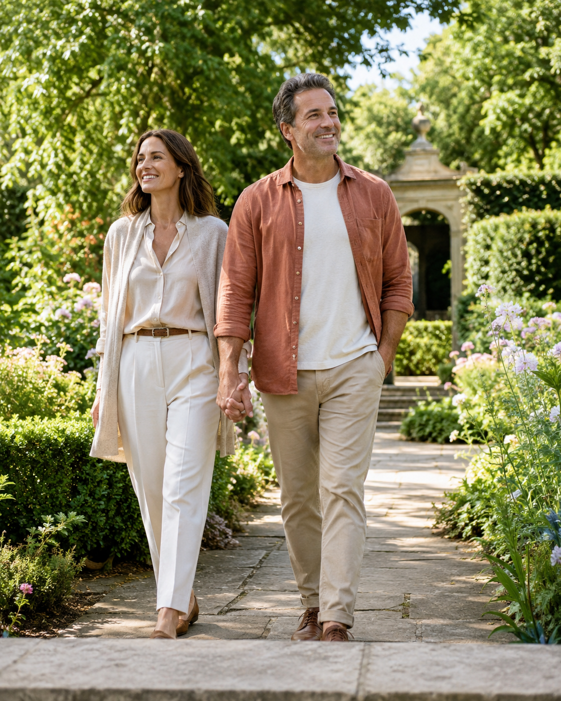 Couple in their 40s holding hands while walking through a sunlit garden, smiling and looking ahead.